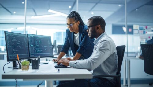 Two software developers collaborate on a coding project in a modern office setting. They work together on a computer, focused on lines of code. Teamwork and technology concept.